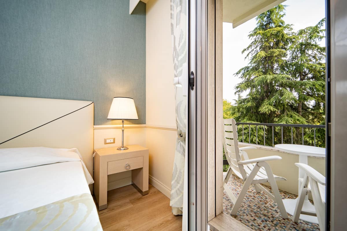 Habitación de hotel con cama, mesita de noche, lámpara y balcón con sillas y vistas a árboles.
