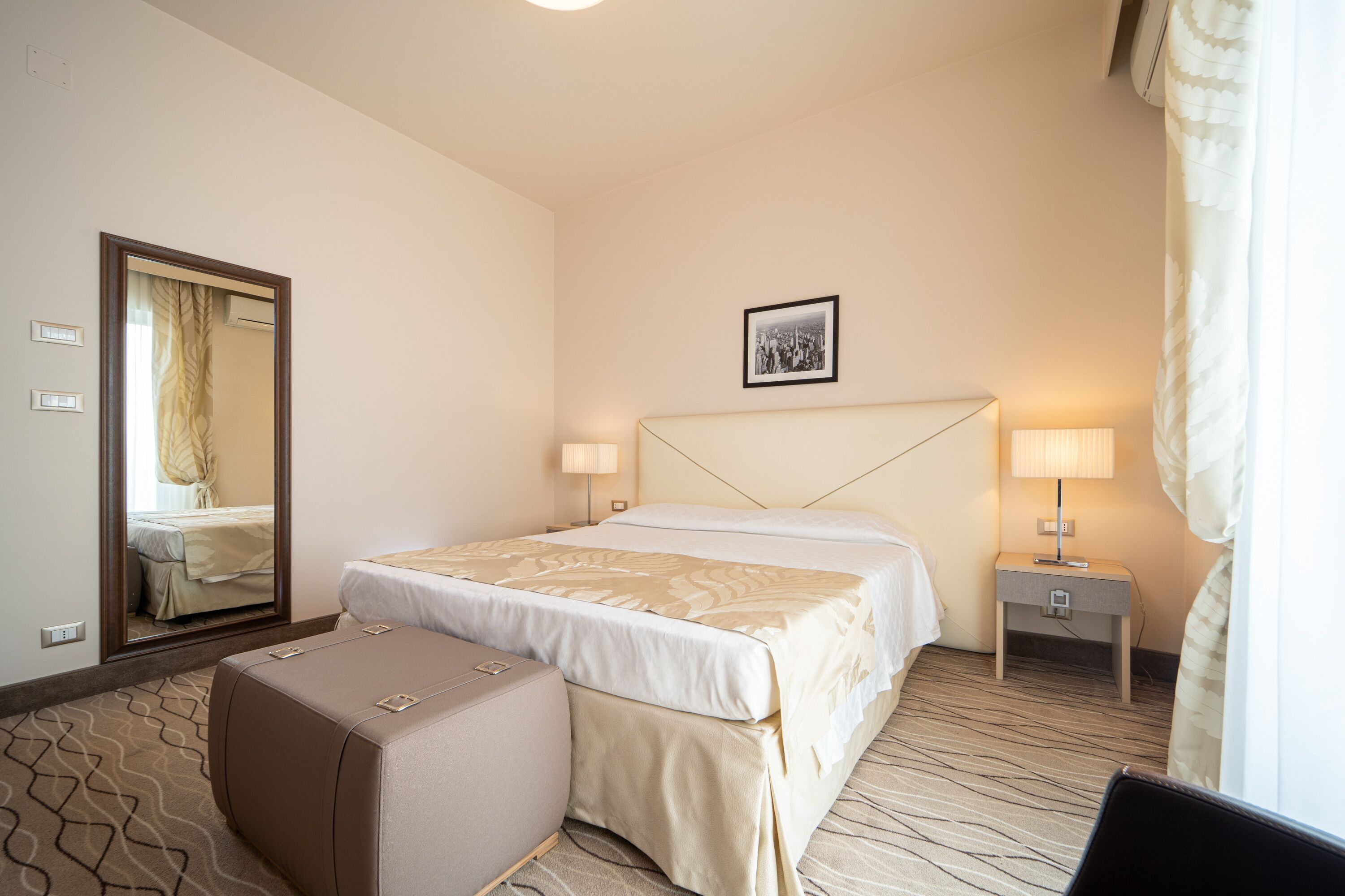 A hotel room with a large bed, a bedside table with a lamp, a small table with a lamp, a suitcase, and a mirror on the wall.