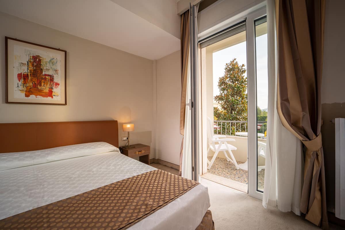 Habitación de hotel con cama, mesita de noche, lámpara, un cuadro en la pared y puertas correderas de cristal que dan a un área exterior con mesa y sillas.