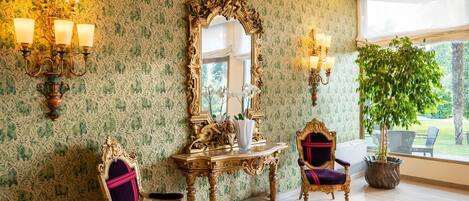 Una habitación con un espejo grande y ornamentado, dos sillas, una consola con un florero y una planta en maceta.
