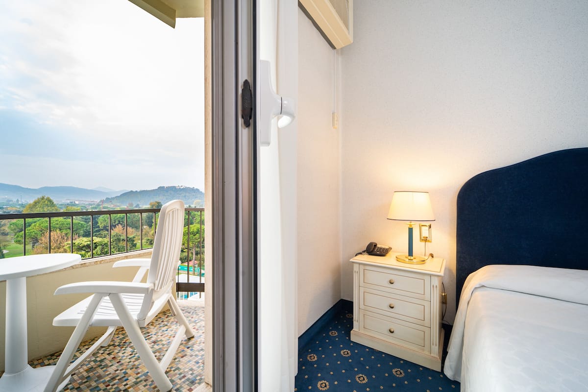 Habitación de hotel con balcón, una cama, una mesita de noche con lámpara y una silla.