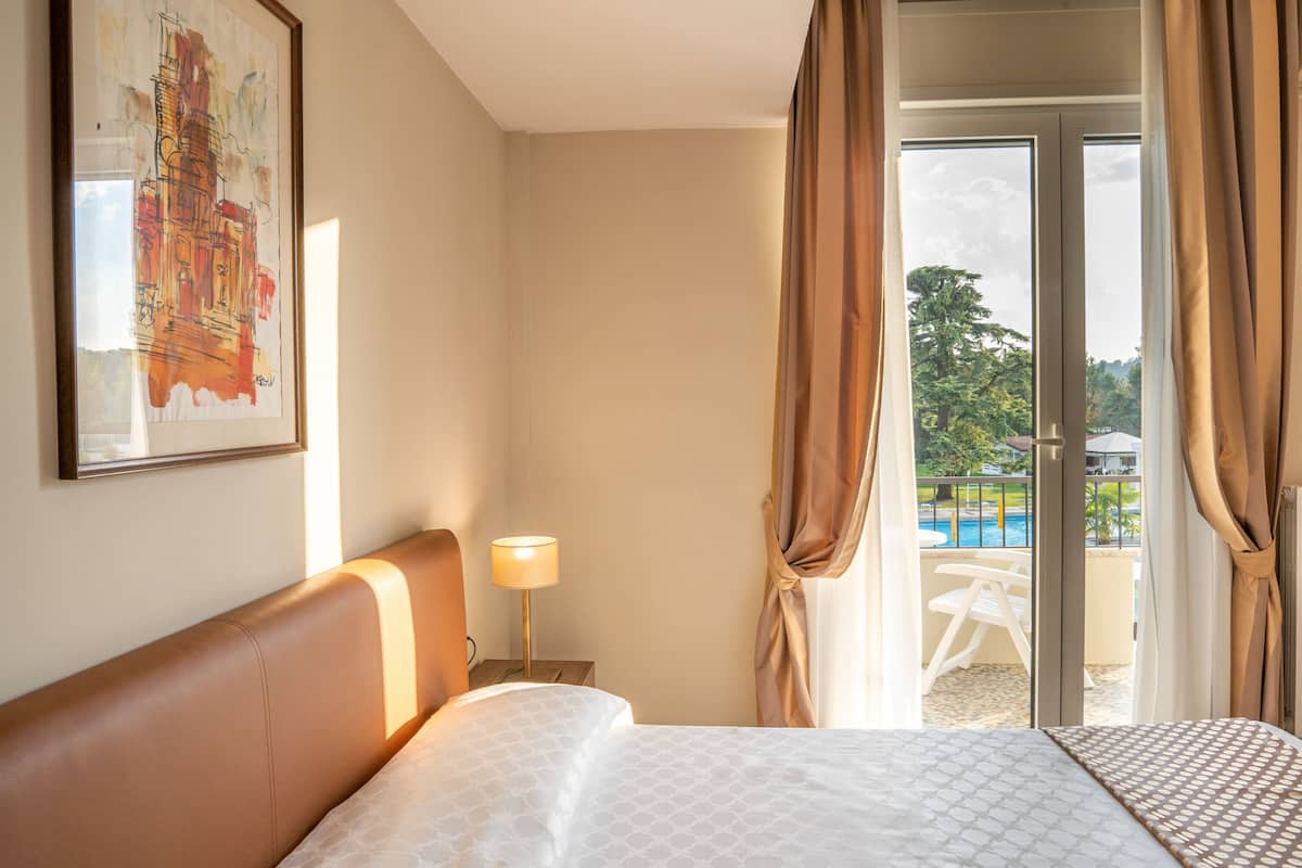 Habitación de hotel con cama, mesita de noche con lámpara, un cuadro en la pared y vistas a una piscina y vegetación por la ventana.