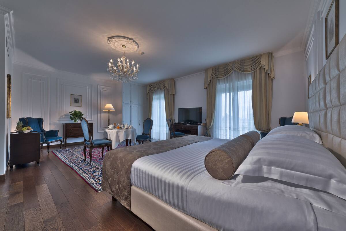 A spacious hotel room with a large bed, a chandelier, a desk with a chair, a television, and a view of the city through the windows.