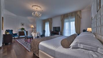 A spacious hotel room with a large bed, a chandelier, a desk with a chair, a television, and a view of the city through the windows.