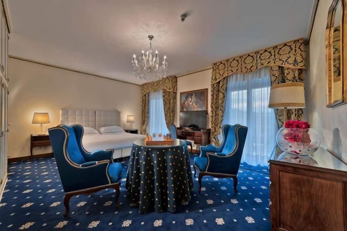 A hotel room with a bed, two blue armchairs, a round table, a chandelier, a dresser, and a mirror.