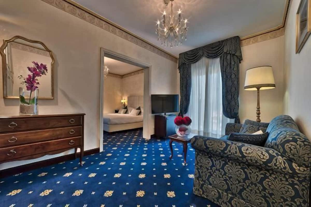 A hotel room with a blue patterned carpet, a sofa, a TV, a dresser, and a chandelier.