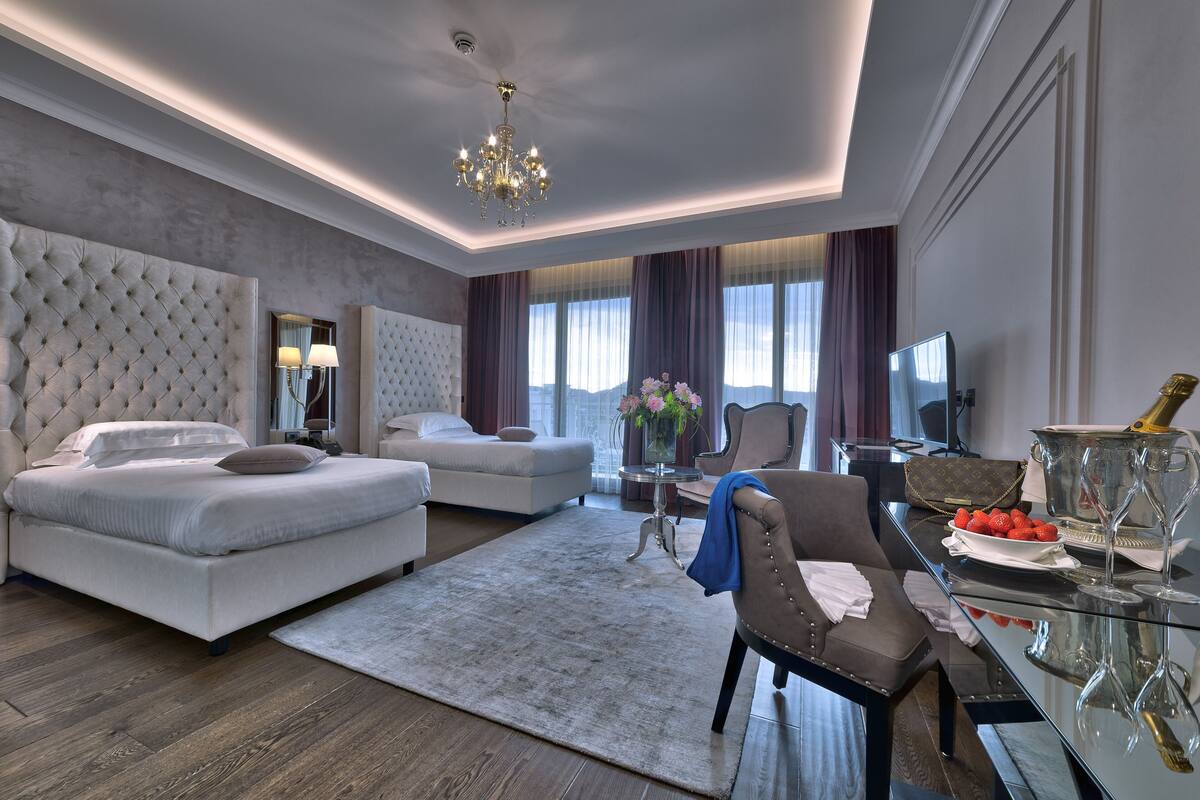 A modern hotel room with a large bed, a sofa, a dining table with a vase of flowers, and a view of the city.