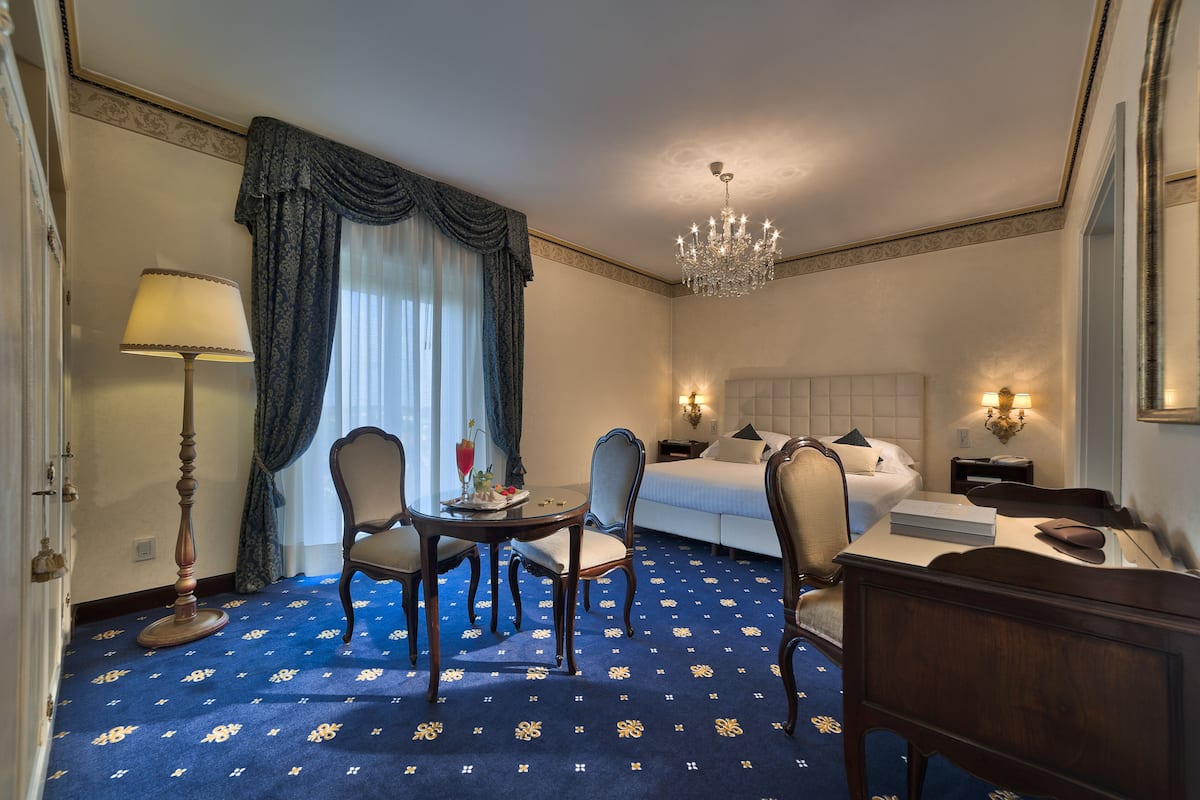 A hotel room with a bed, two chairs, a table, a lamp, and a chandelier.
