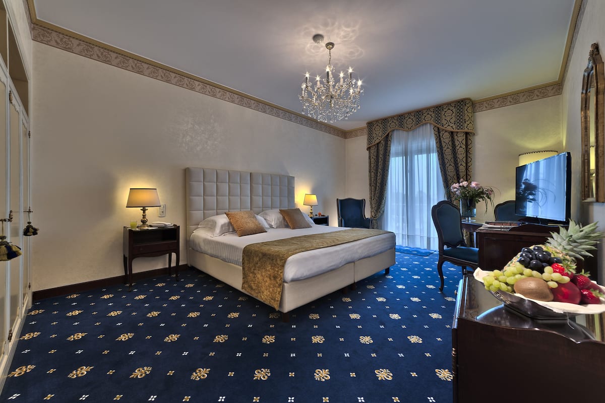 A hotel room with a large bed, two bedside tables with lamps, a chandelier, a desk with a computer, and a fruit bowl on a dresser.