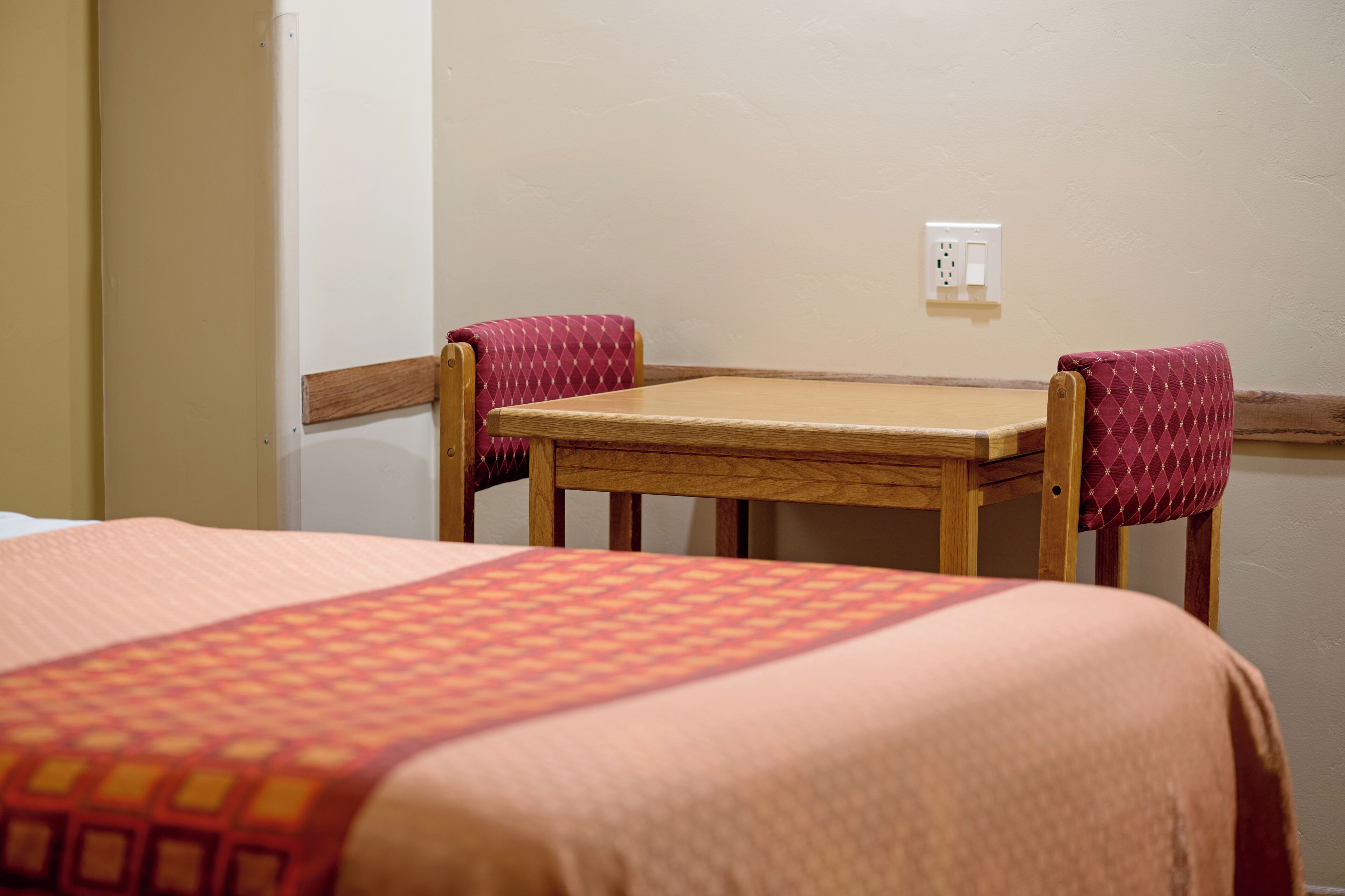 Une chambre d’hôtel avec un lit simple, une table en bois, deux chaises rouges avec des coussins et une prise électrique au mur.