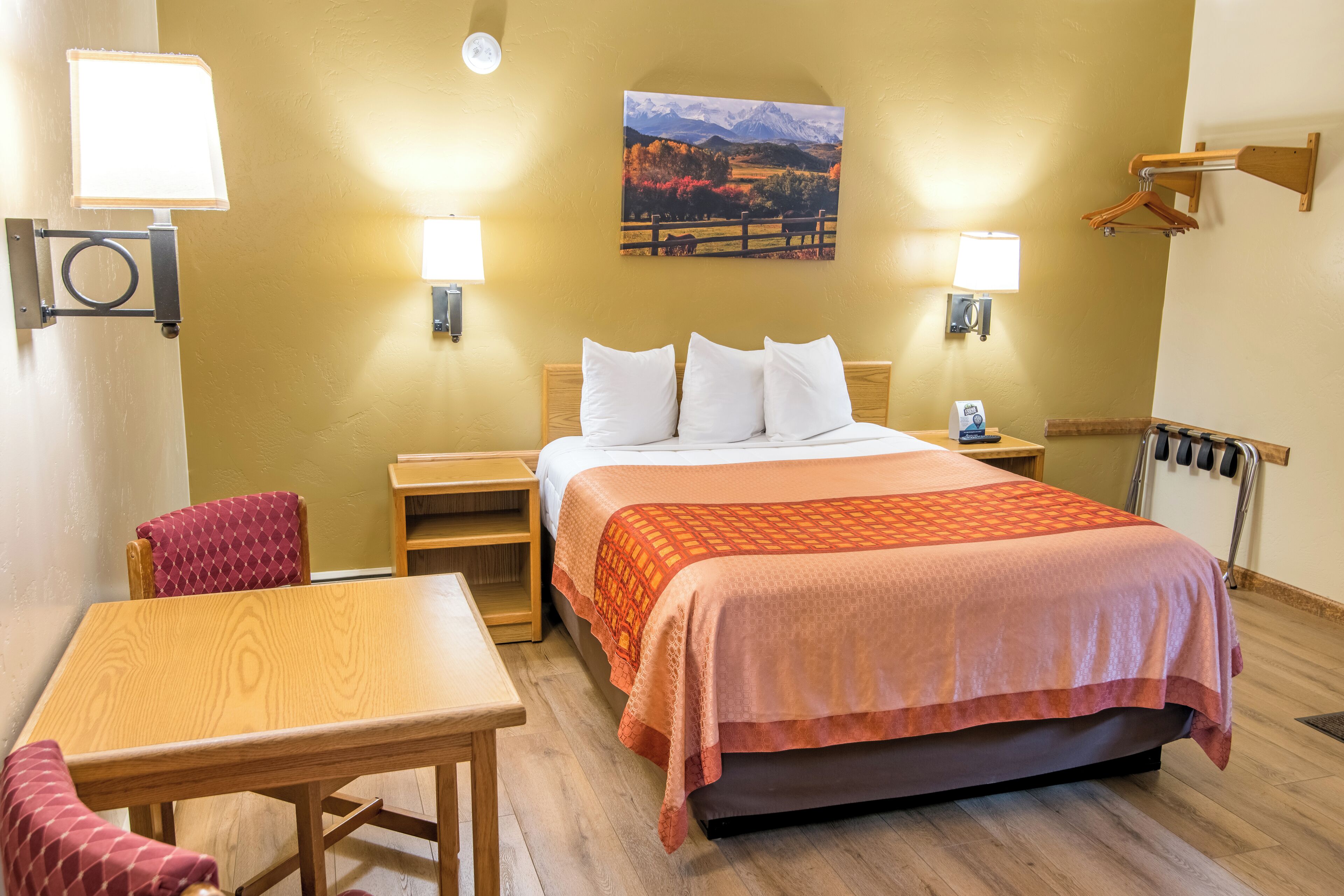 A hotel room with a bed, a wooden table, a chair, two bedside lamps, a wall-mounted heater, and a painting of a landscape.