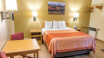 A hotel room with a bed, a wooden table, a chair, two bedside lamps, a wall-mounted heater, and a painting of a landscape.