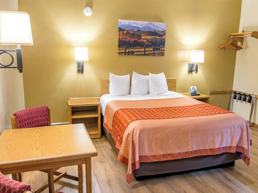 A hotel room with a bed, a wooden table, a chair, two bedside lamps, a wall-mounted heater, and a painting of a landscape.