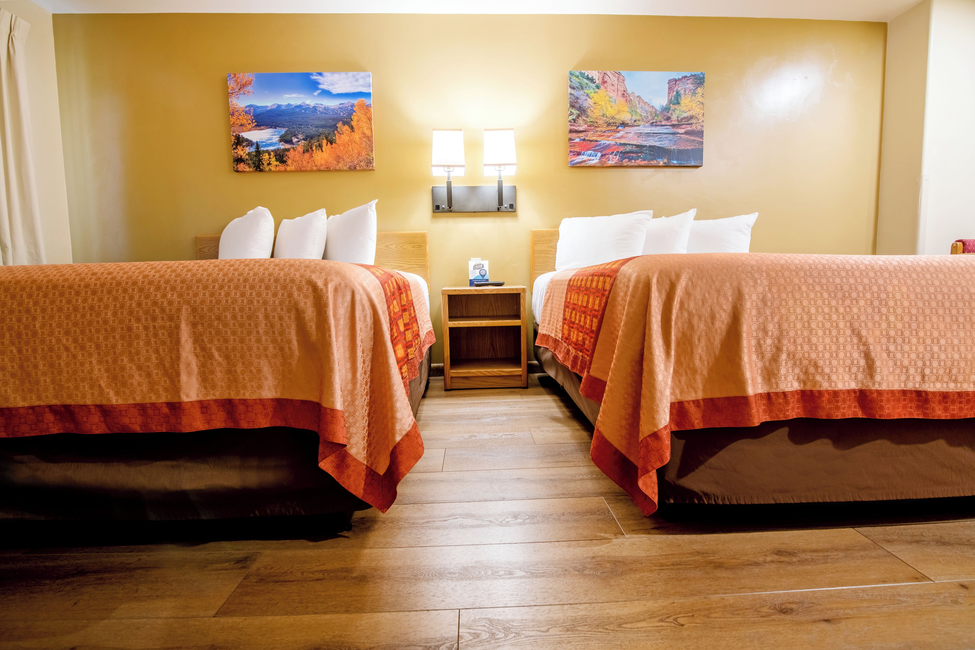 Habitación de hotel con dos camas, una mesita de noche con lámpara y dos cuadros en la pared.