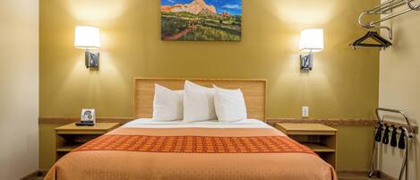 A hotel room with a bed, two bedside lamps, a painting of a mountain landscape, and a coat rack with hangers.