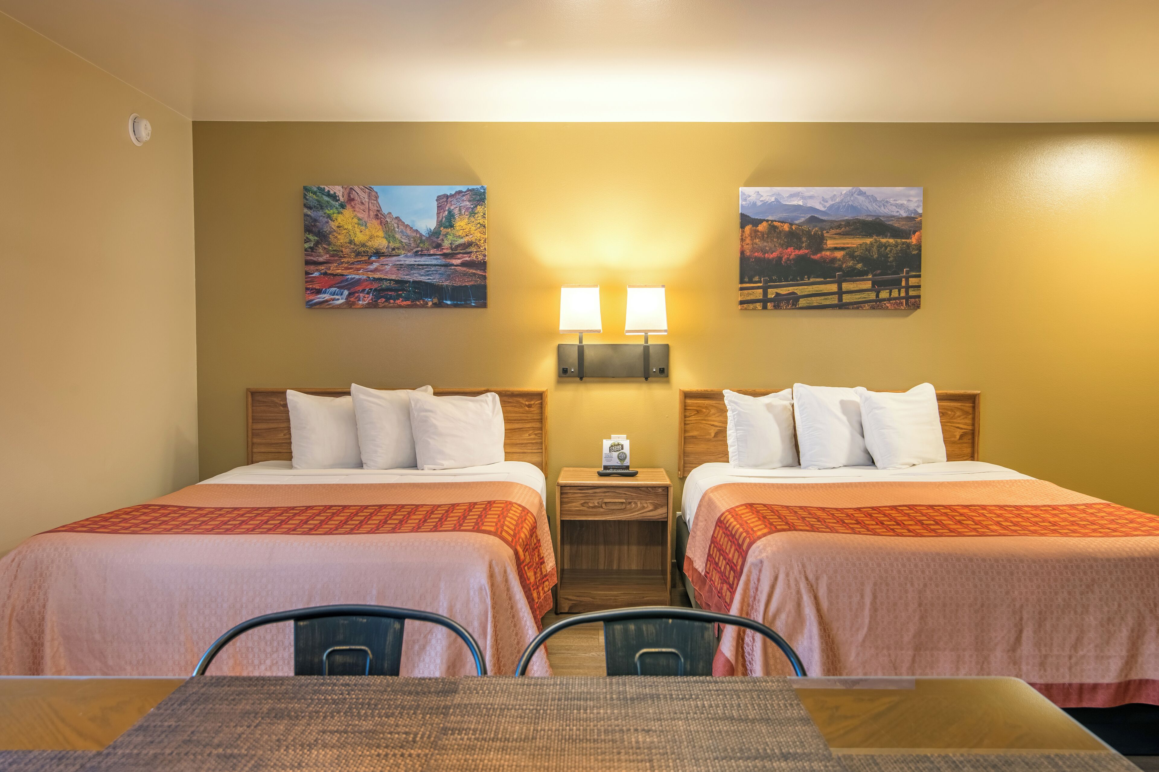 A hotel room with two beds, a small table, and two chairs. There are two wall-mounted lamps and two paintings above the beds.