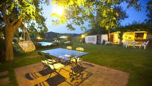 Een terras buiten met een glazen tafel en stoelen, omgeven door een verzorgd gazon en bomen.
