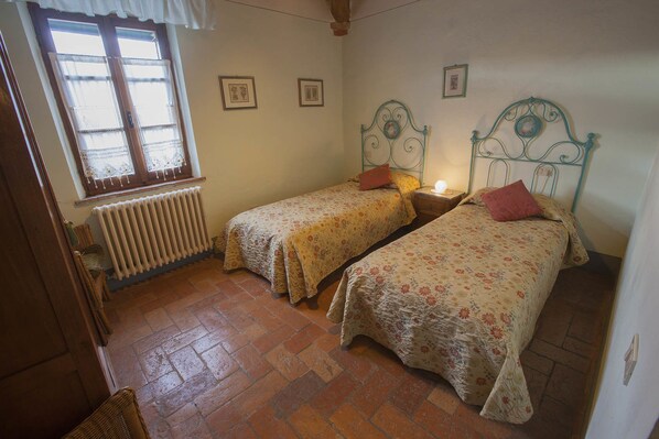 Une chambre avec deux lits, un radiateur, une fenêtre ornée de rideaux en dentelle et une chaise en bois.