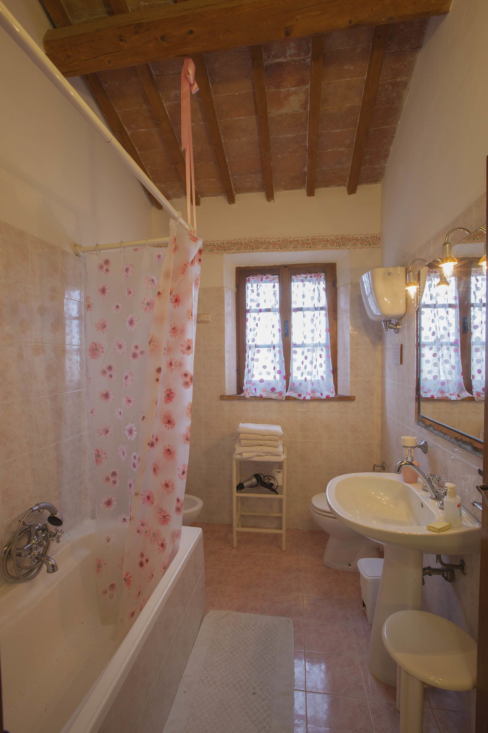 A bathroom with a bathtub, sink, and toilet. There is a window with curtains and a wooden ceiling.