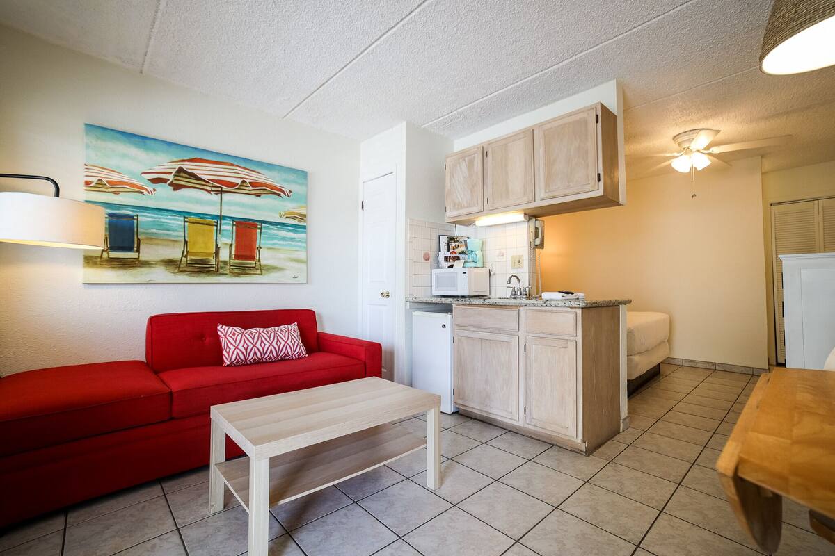A compact living space with a kitchenette, a red sofa, a small table, and a beach-themed painting on the wall.