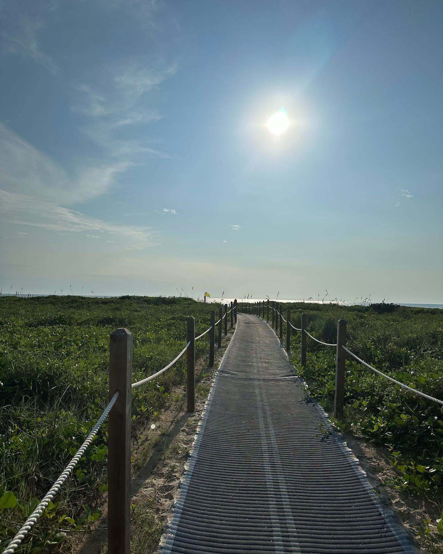 Un sendero que lleva a una playa con cielo despejado y el sol brillando.