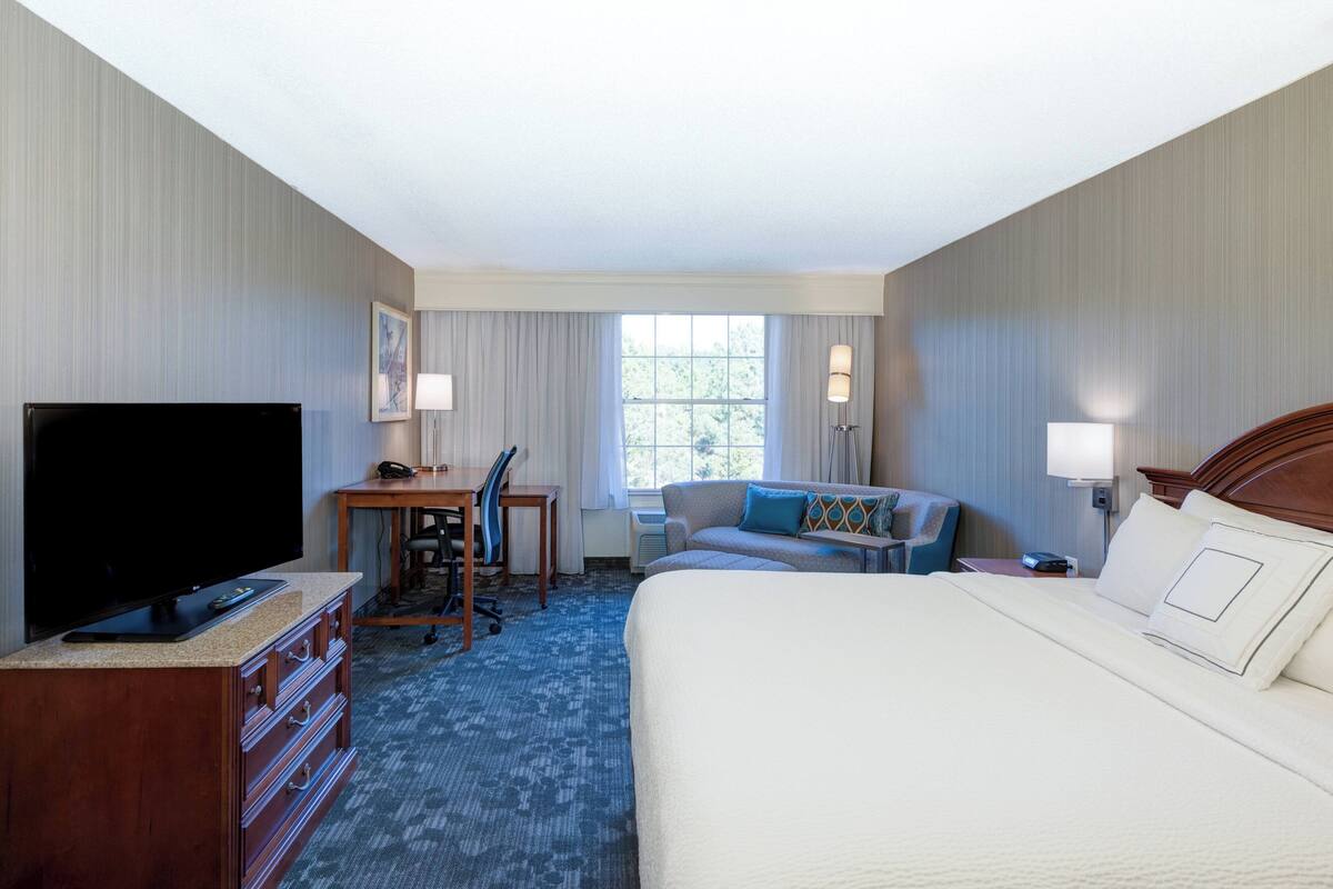 Quarto de hotel com uma cama grande, uma escrivaninha com cadeira, televisão e janela com cortinas.