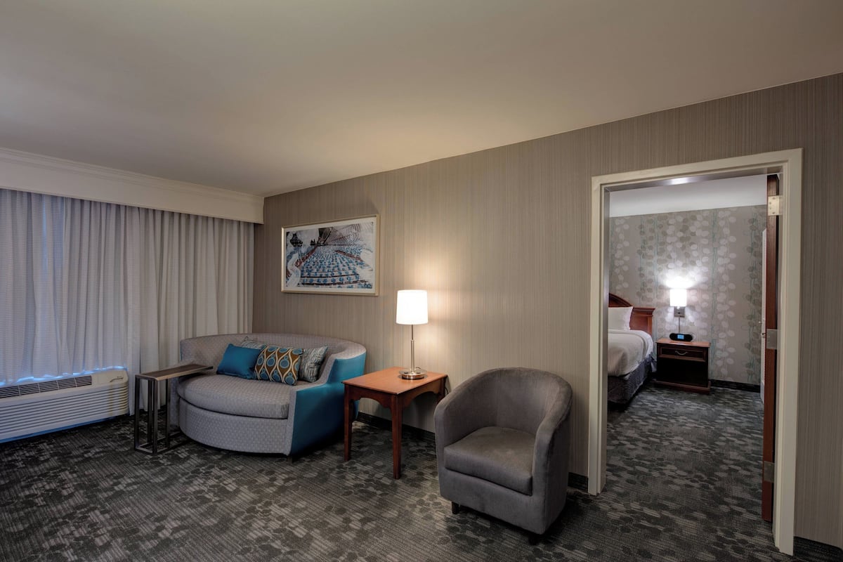 Quarto de hotel com sofá cinzento, uma poltrona pequena, mesa lateral com abajur, pintura na parede e cama com mesa de cabeceira.