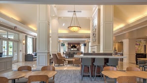 Un amplio hall de hotel con una barra central, zonas de estar y vistas a la cocina.