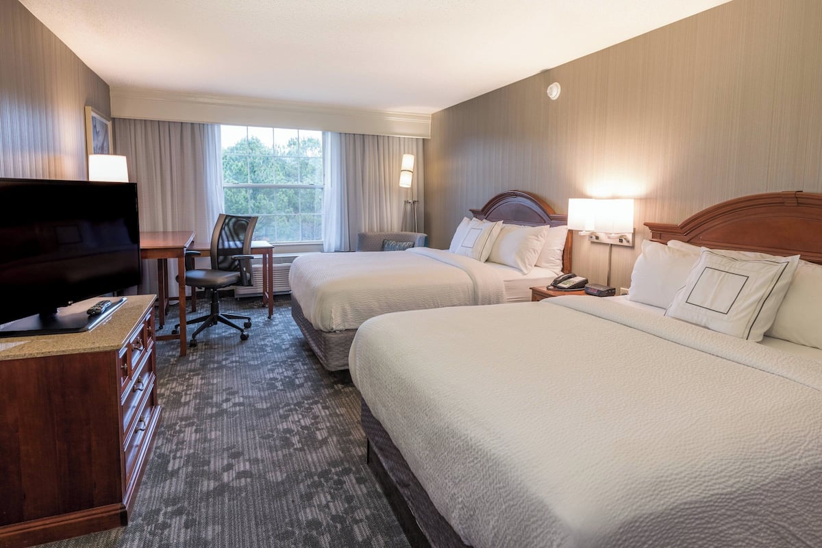 Quarto de hotel com duas camas, uma escrivaninha com televisão e vista para árvores pela janela.
