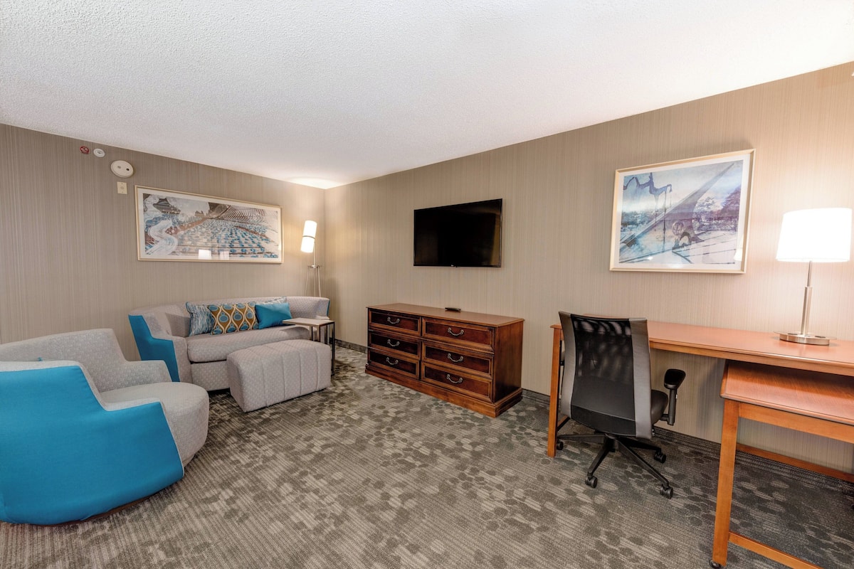 Quarto de hotel com sofá, escrivaninha, cadeira, cômoda e duas televisões fixadas na parede.