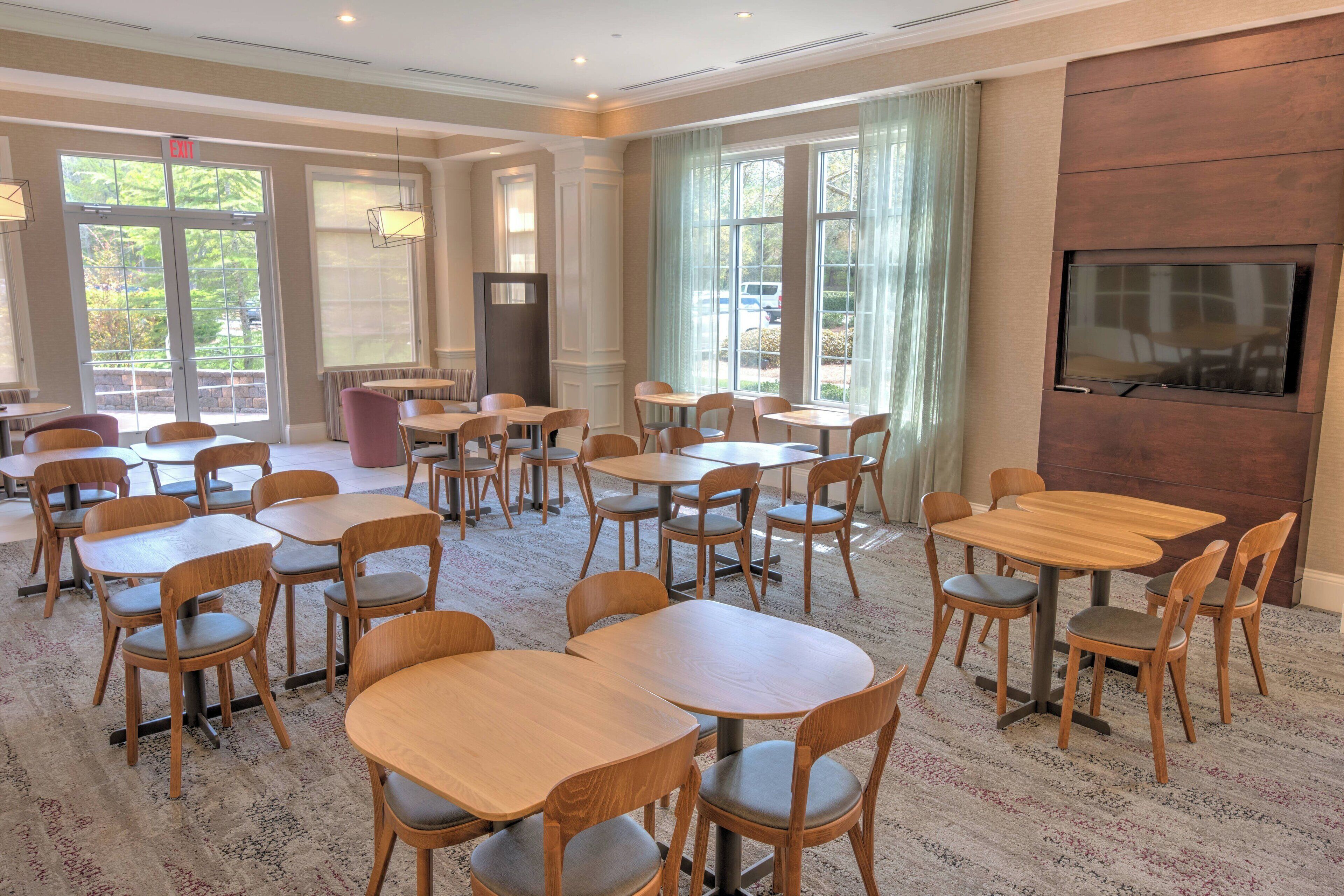 A hotel dining area with multiple tables and chairs, a large TV screen on the wall, and a view of the outdoors through large windows.