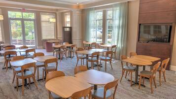 A hotel dining area with multiple tables and chairs, a large TV screen on the wall, and a view of the outdoors through large windows.