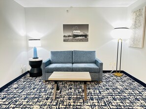 A room with a blue sofa, a small wooden table, a black floor lamp, and a wall-mounted picture. - Best Western Plus Omaha Airport Inn (Carter Lake)