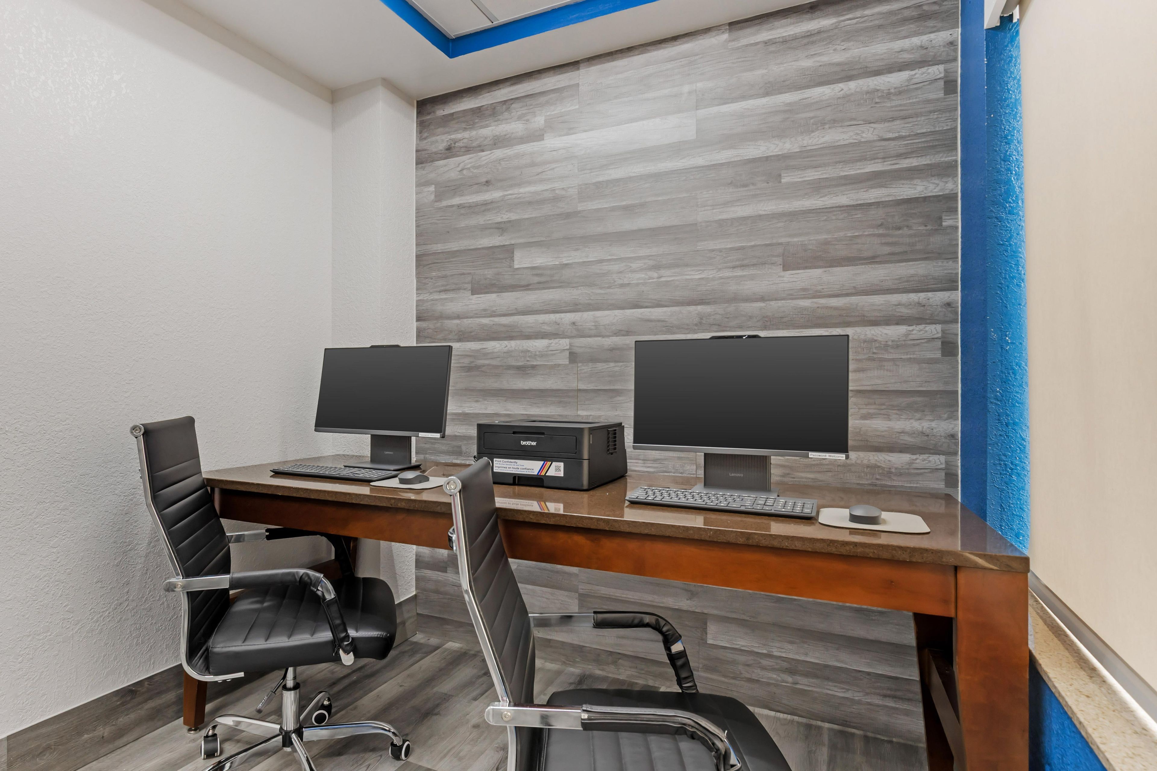 A modern office space with two desks, each with a computer monitor, keyboard, and mouse. There are two black office chairs with wheels. The room has a wooden floor and a grey wall with a blue accent.