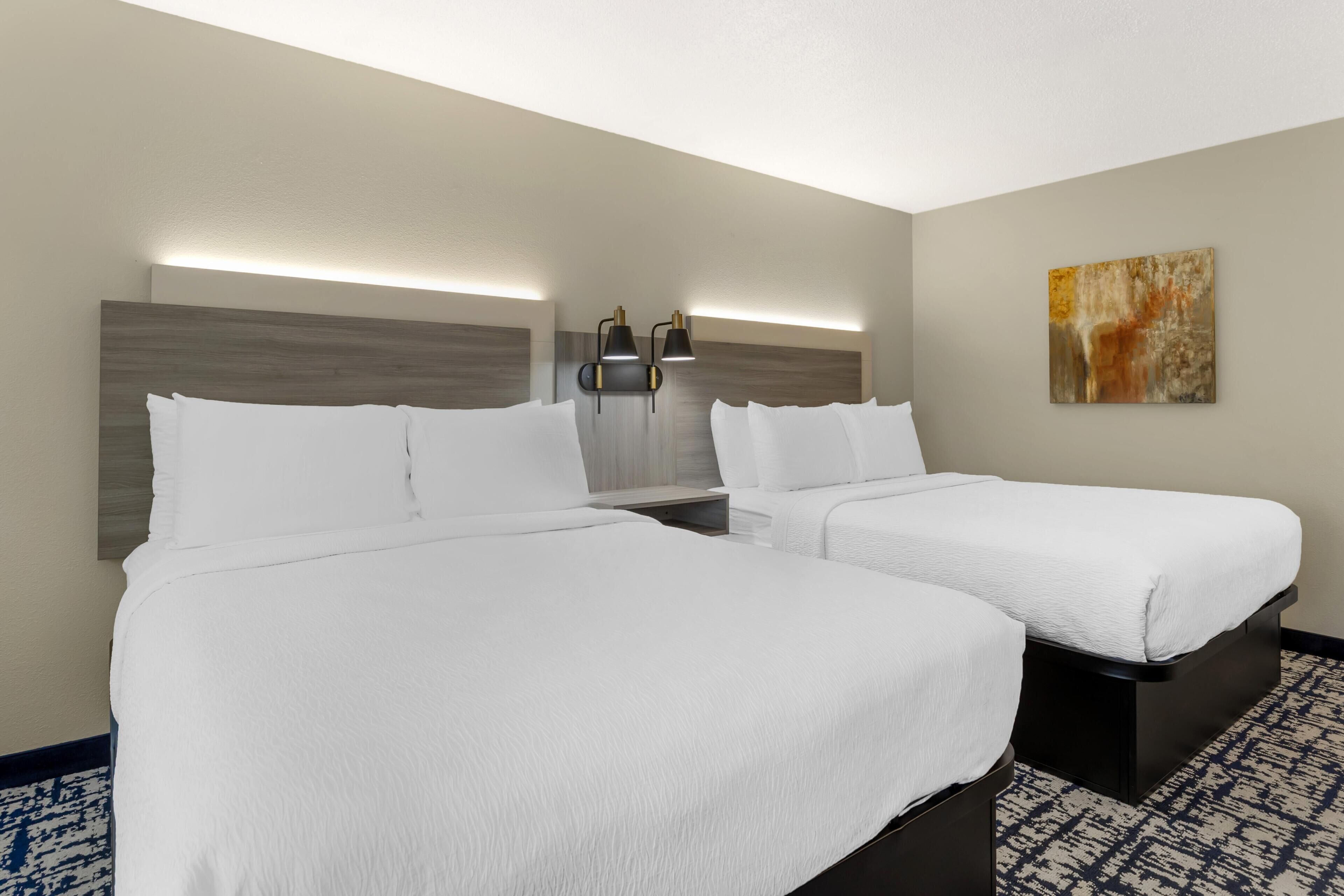 A hotel room with two beds, a nightstand, a wall-mounted lamp, and a painting.