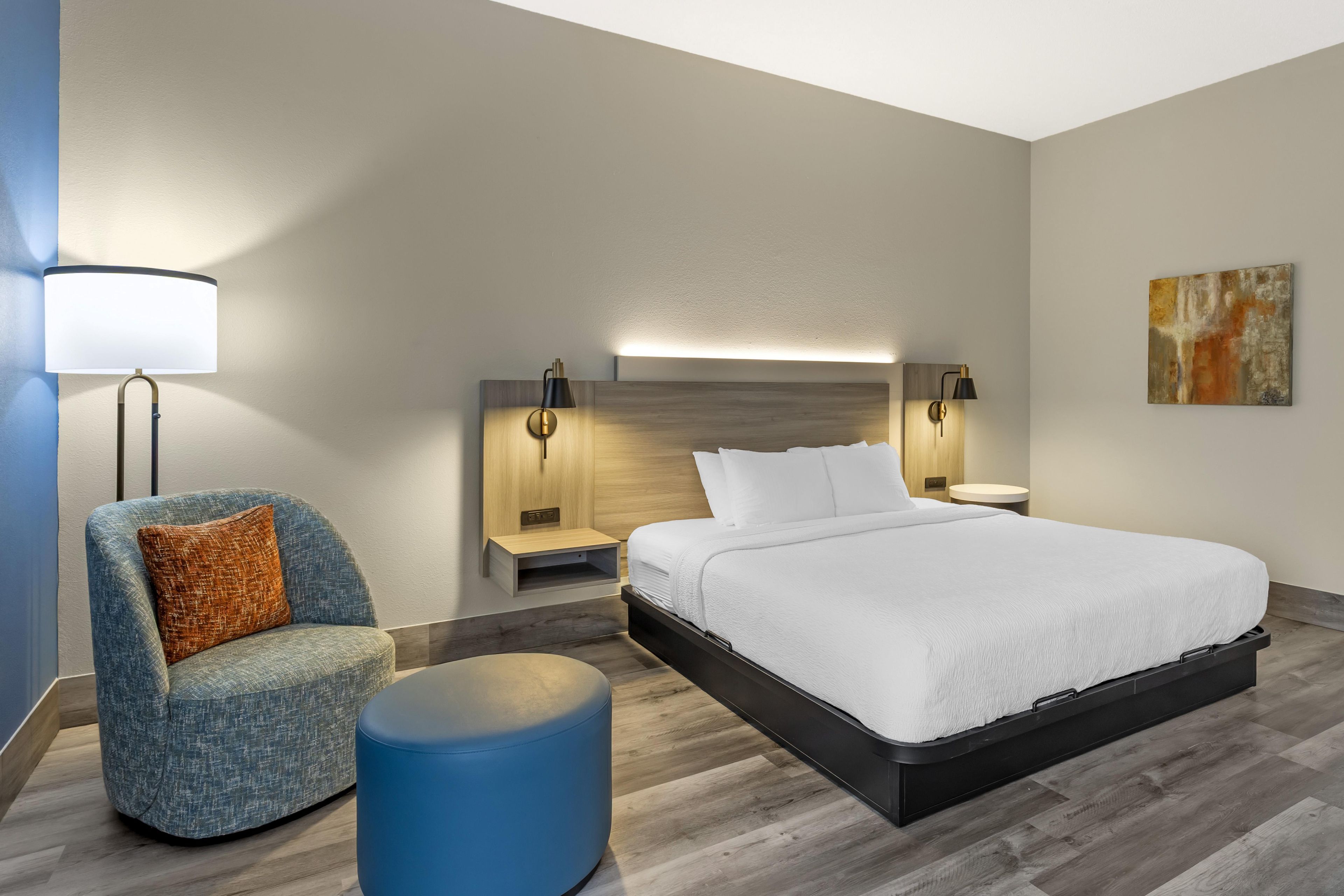 Ein modernes Hotelzimmer mit Bett, einer Sitzecke mit Sessel, einem Nachttisch und einer Wandlampe.