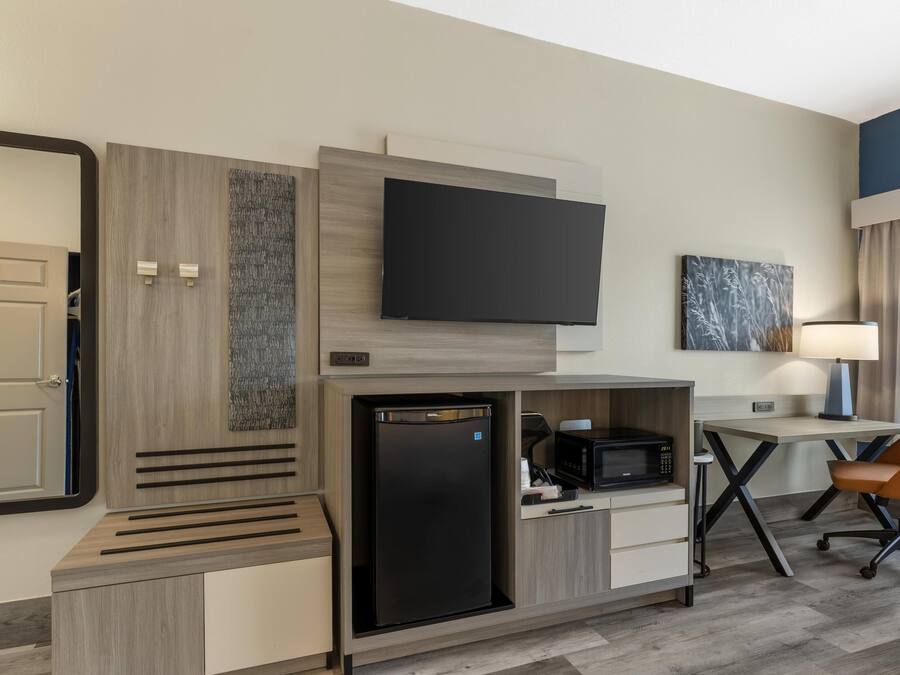 A compact hotel room with a flat-screen TV, a mini-fridge, a microwave, and a seating area with a chair and table.