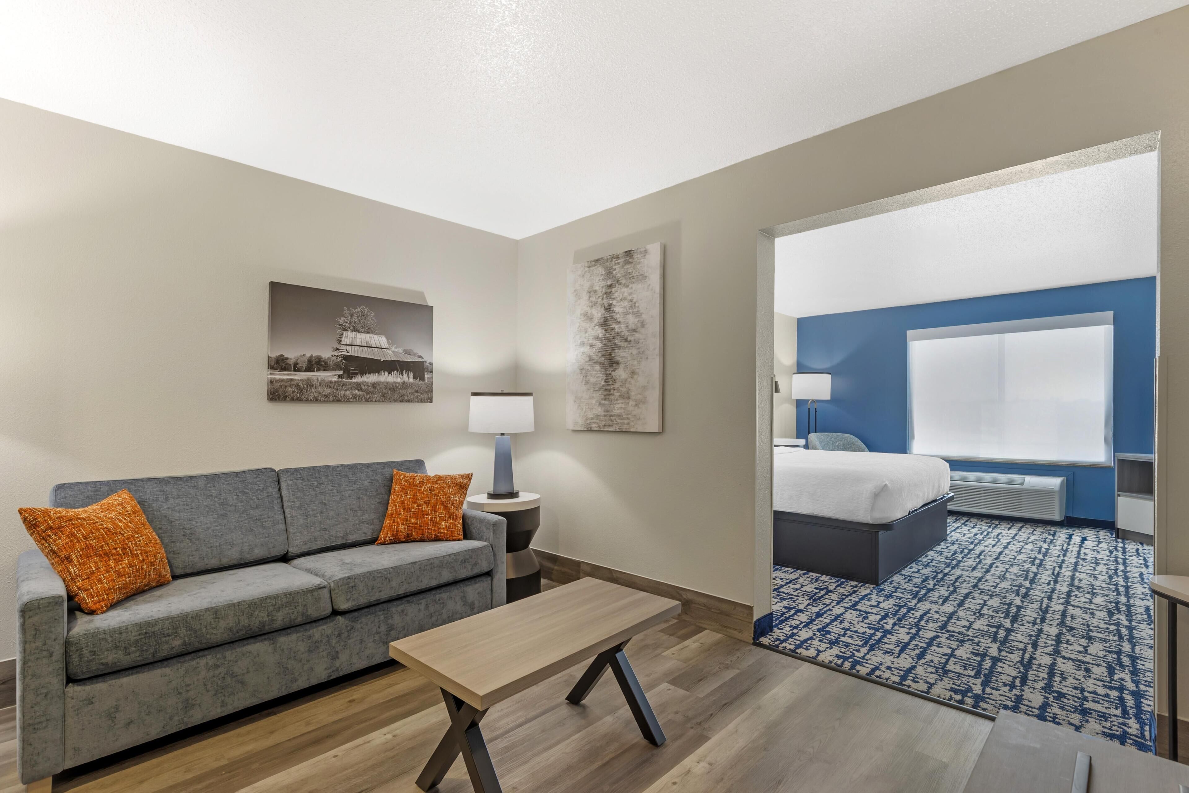 A hotel room with a gray sofa, a wooden coffee table, a bed with a blue headboard, and a large window.