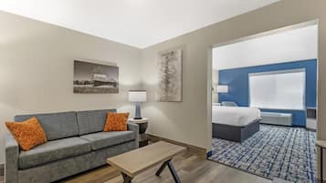 A hotel room with a gray sofa, a wooden coffee table, a bed with a blue headboard, and a large window.
