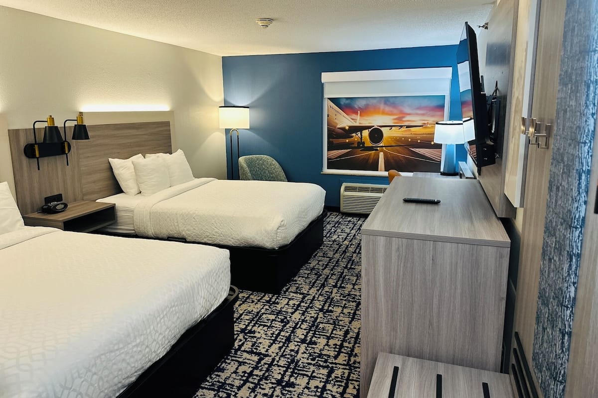 A hotel room with two beds, a desk, a chair, a TV, and a painting of an airplane on the wall.