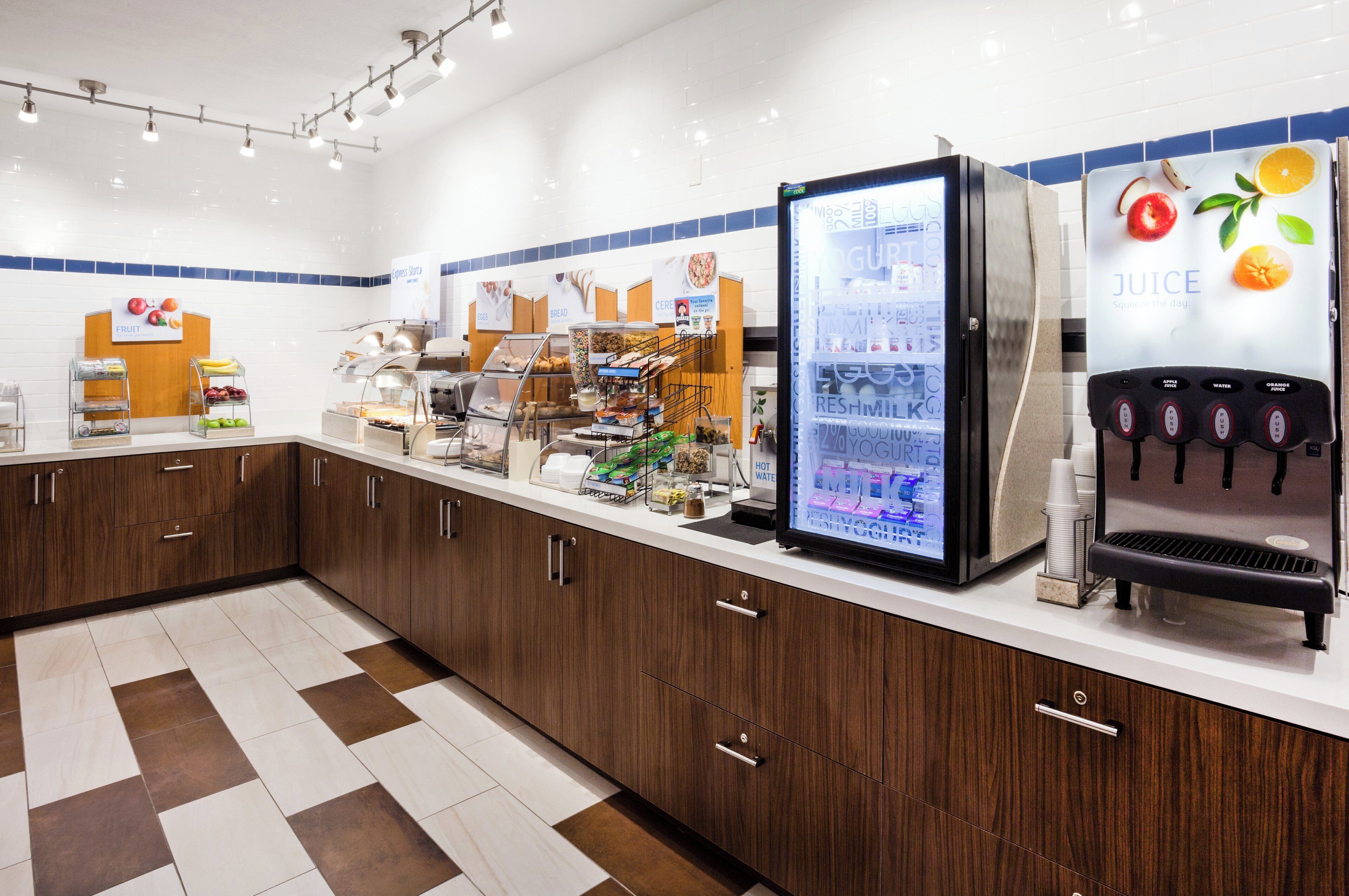 A self-service breakfast area with a refrigerated display, a juice dispenser, and various food items.