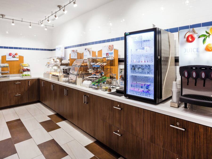 A self-service breakfast area with a refrigerated display, a juice dispenser, and various food items.