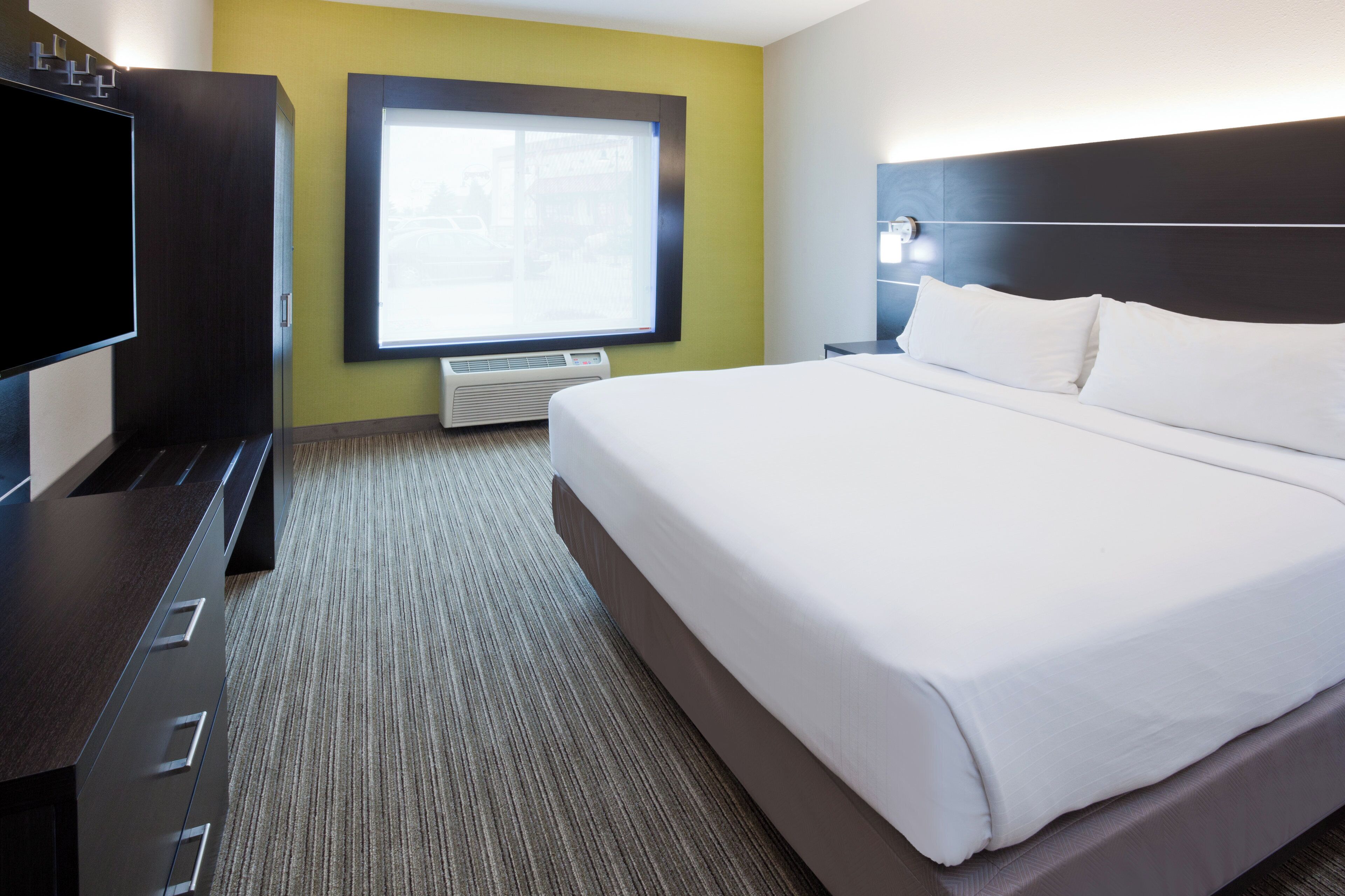 A hotel room with a large bed, a flat-screen TV, a window with blinds, and a wall-mounted heater.