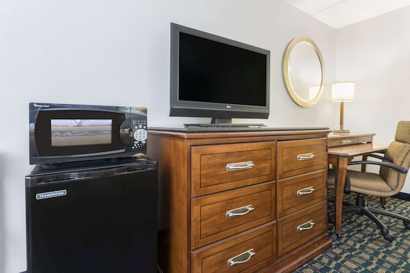 A hotel room with a minibar, a flat-screen TV, a wooden dresser with drawers, a chair, a desk, and a lamp.