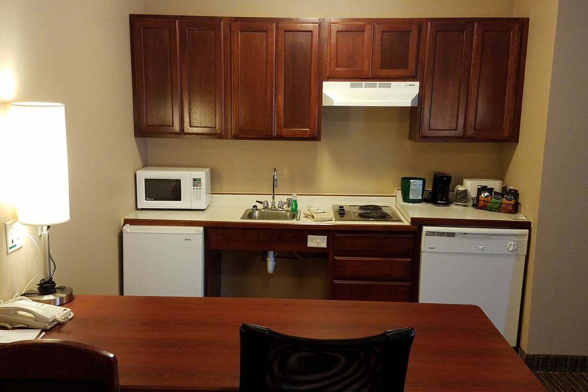 Cocina compacta con armarios de madera, un microondas, un lavabo y una estufa. Hay una mesa con una silla adelante.