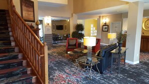 A hotel lobby with a staircase, a reception desk, a seating area with chairs, and a dining area. - GrandStay Residential Suites - Eau Claire (Eau Claire)