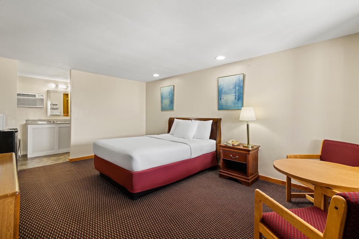 A hotel room with a bed, a desk, a chair, a table, a lamp, and a painting on the wall.