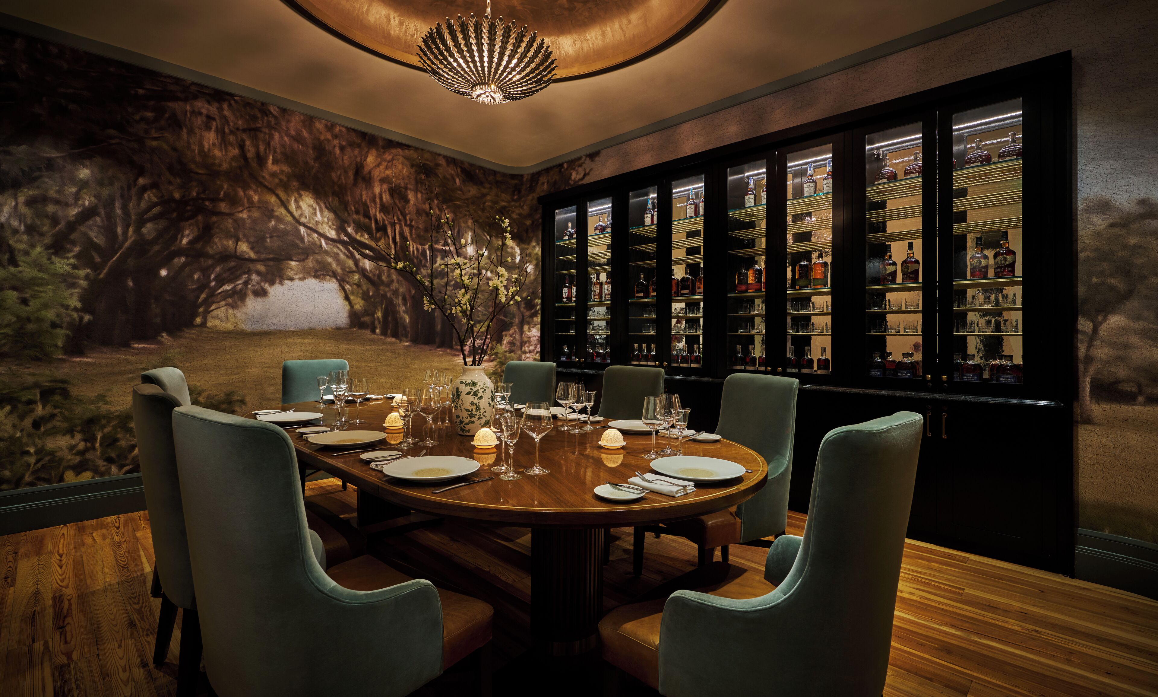 Un comedor con una gran mesa redonda dispuesta para seis personas, rodeada por seis sillas. Un mural en la pared representa una escena de bosque, y se aprecia una barra con estantes llenos de botellas de licor al fondo.