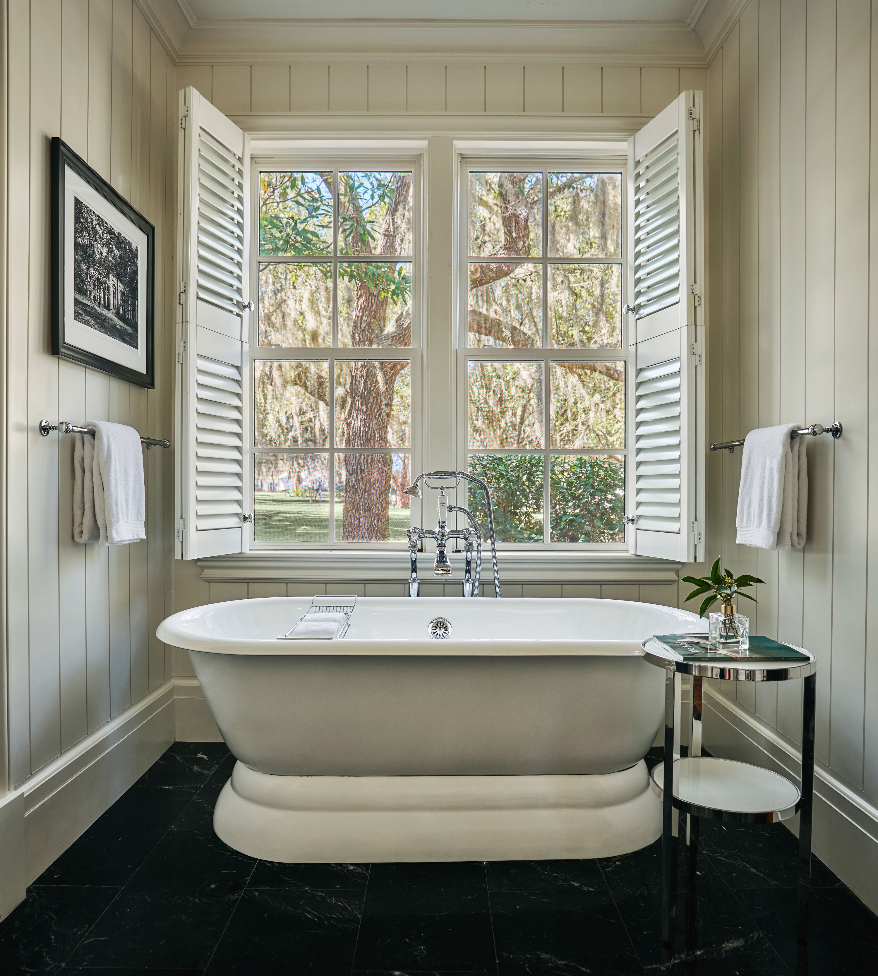 A bathroom with a freestanding bathtub, a window with shutters, and a framed picture on the wall.