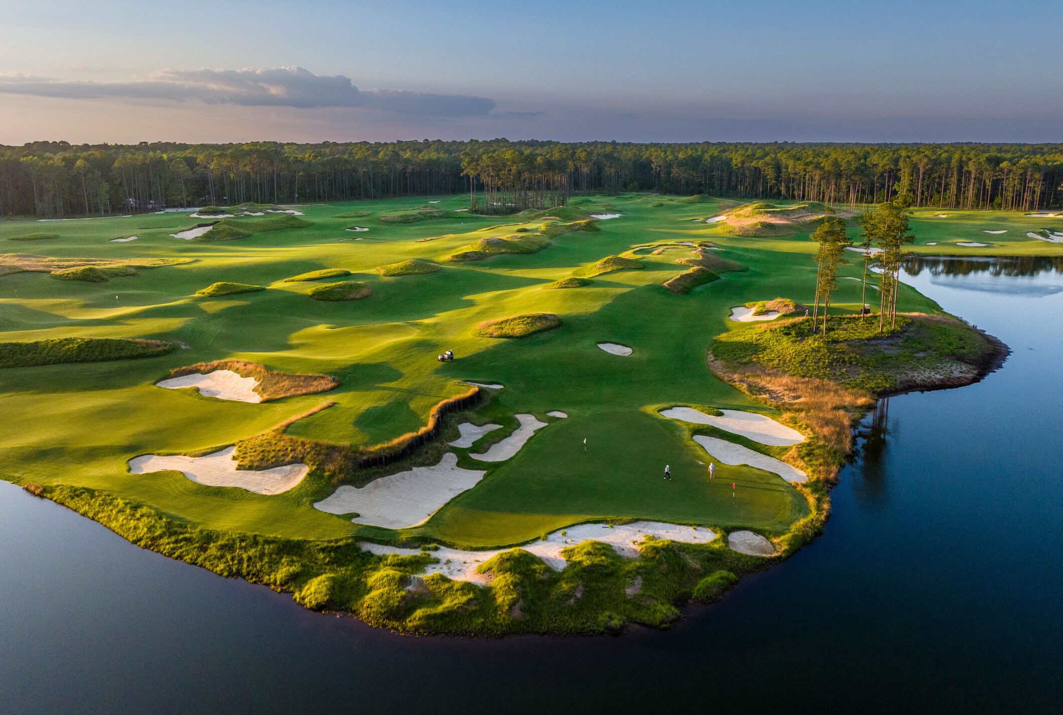 Un campo de golf con búnkeres de arena, obstáculos acuáticos y un bosque al fondo.